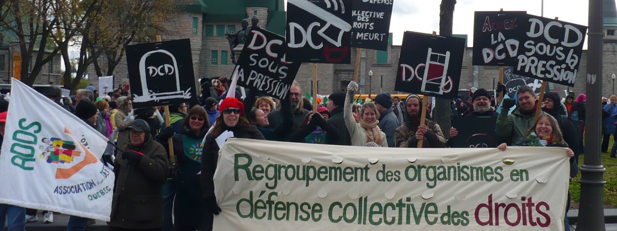 Photo : employé-es des organismes qui manifestent, très souriants et enjoués. Belle bannière : Regroupement des organismes en défense collective des droits ; ce dernier moi est en rouge. Plusieurs pancartes noires : DCD sous pression, image de pierre tombale. On voit le Manège militaire derrière.